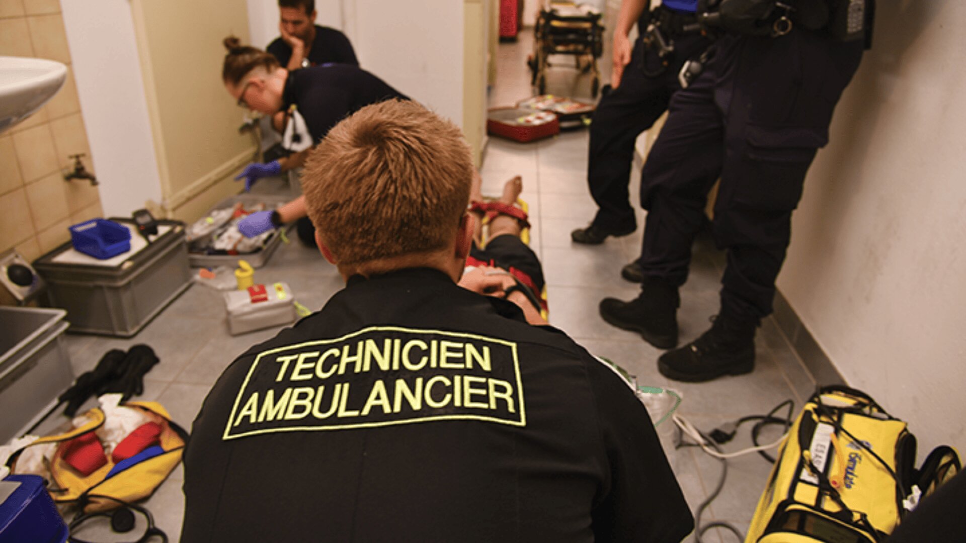Des ambulanciers et des intervenants d'urgence s'occupent d'une personne allongée sur le sol dans un couloir étroit, avec du matériel médical et des fournitures éparpillés. Au dos de l'un des uniformes, on peut lire "TECHNICIEN AMBULANCIER".