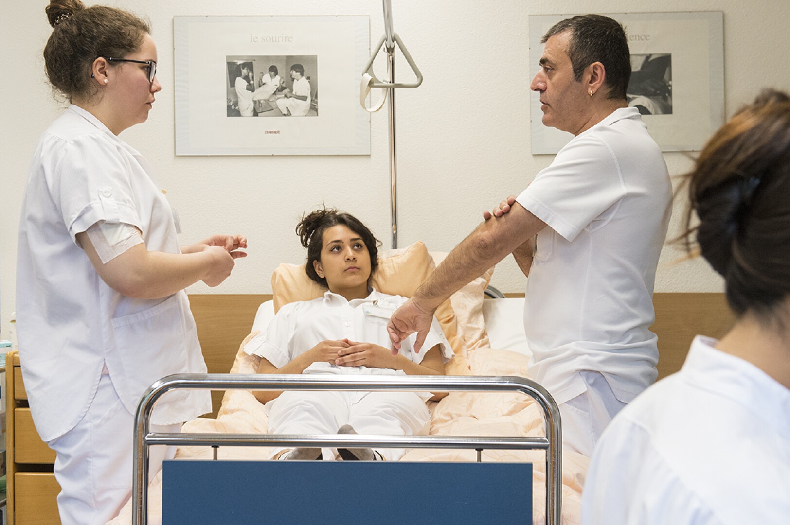 Deux professionnels de santé discutent à côté d'une femme allongée dans un lit d'hôpital, tandis qu'une autre personne est assise au premier plan. Le décor est une chambre d'hôpital avec du matériel médical et des photos encadrées au mur.