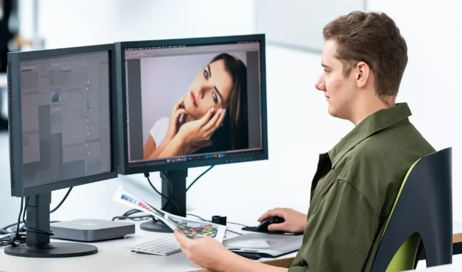 Une personne est assise à un bureau, utilisant un ordinateur avec deux moniteurs, éditant le portrait d'une femme sur un écran tout en tenant un magazine ou des documents imprimés dans l'autre main.