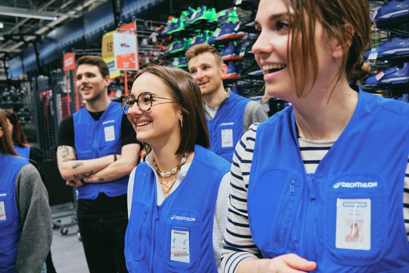 Un groupe d'employés souriants portant des gilets bleus et des badges se tient ensemble à l'intérieur d'un magasin de sport, entouré d'étagères de chaussures de sport colorées à l'arrière-plan.