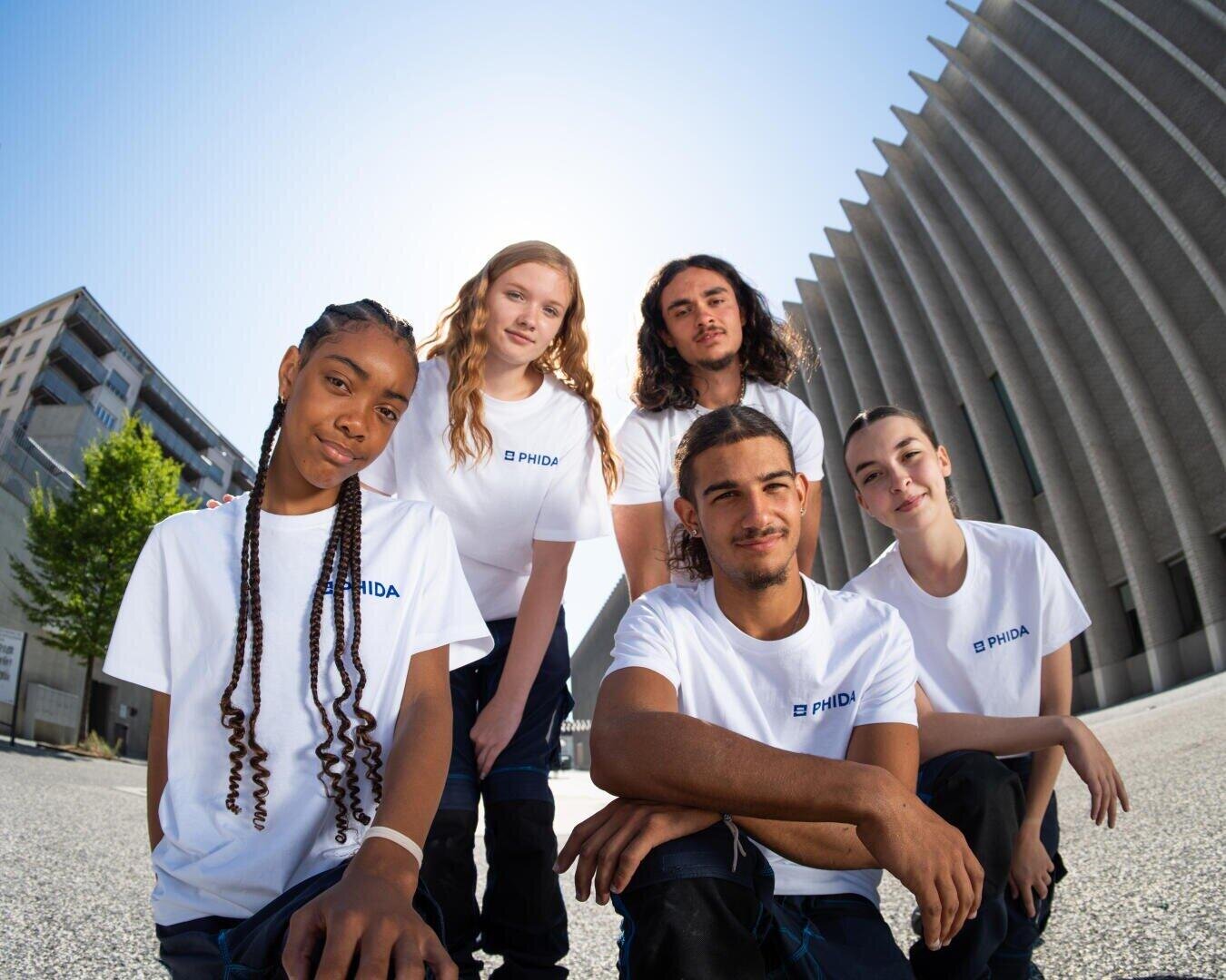 Cinq jeunes adultes portant des t-shirts PHIDA blancs assortis posent ensemble en plein air dans un environnement urbain, souriant avec assurance. L'arrière-plan comprend des bâtiments modernes et un ciel bleu.