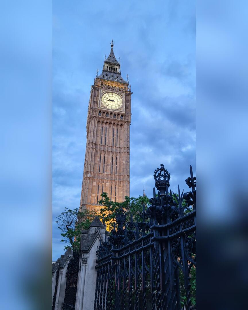 Tour d'horloge Big Ben à Londres, vue d'en bas au crépuscule, avec une clôture décorative en fer noir et un feuillage vert au premier plan, et un ciel bleu nuageux à l'arrière-plan.
