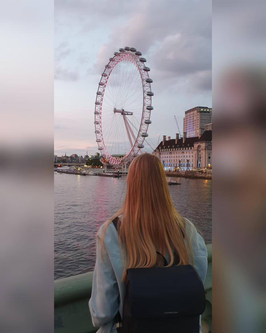 Une personne aux longs cheveux blonds et au sac à dos sombre se tient face à la Tamise, regardant la grande roue London Eye pendant le coucher du soleil. Le ciel est partiellement nuageux et des bâtiments urbains bordent la rivière.