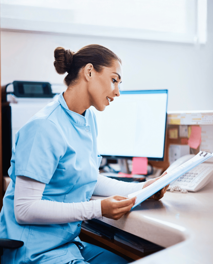 Une infirmière en blouse bleue est assise à un bureau devant un ordinateur et lit des documents. L'espace de travail comprend des fournitures de bureau, un téléphone et des papiers, la lumière naturelle provenant d'une fenêtre à l'arrière-plan.