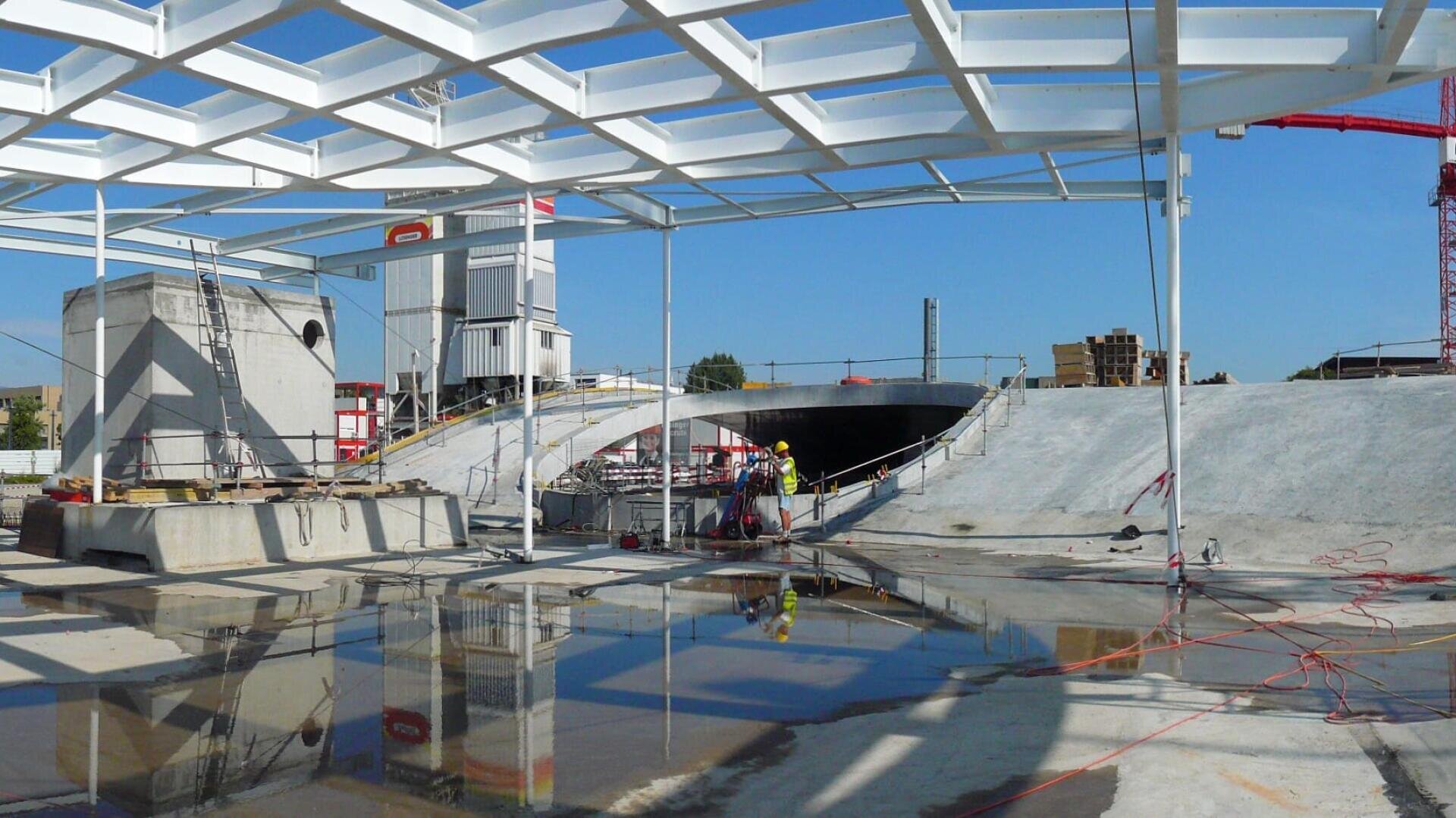 Chantier de construction comprenant une grande structure en béton, une charpente métallique partielle au-dessus de la tête et des ouvriers en tenue de sécurité. Il y a des flaques d'eau sur le sol et des matériaux de construction éparpillés. Le ciel est clair et bleu.