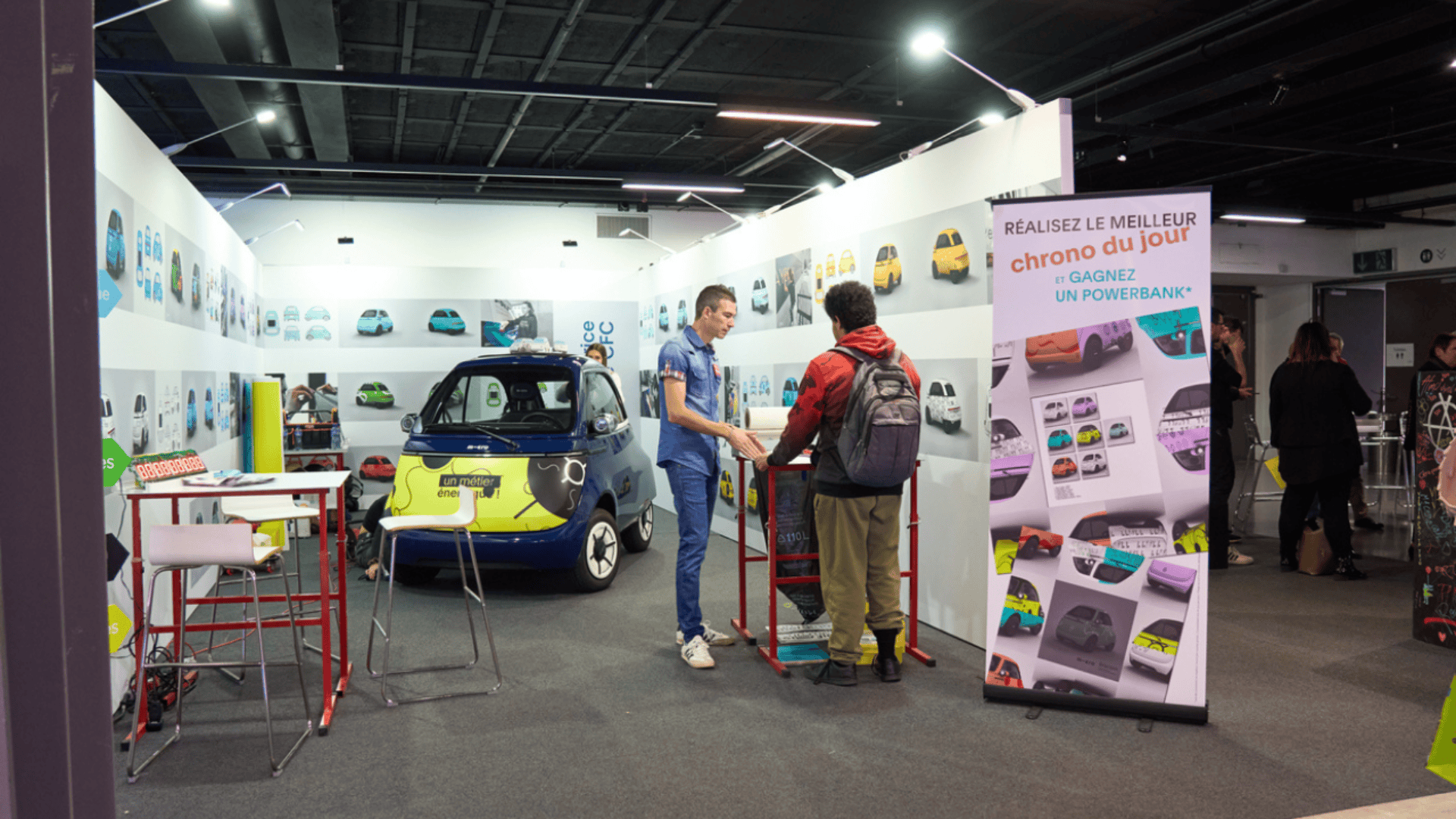 Une petite voiture électrique est exposée dans un stand d'événement intérieur. Deux personnes se tiennent à un comptoir et discutent. Une bannière, des chaises et des graphiques colorés de voitures décorent le stand. D'autres participants sont visibles à l'arrière-plan.
