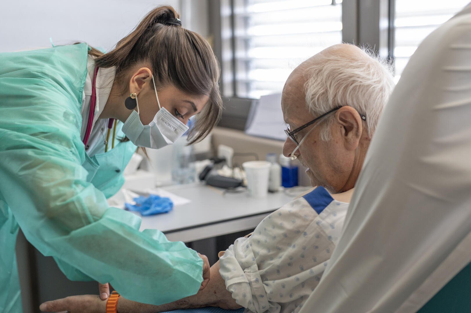 Un agent de santé portant des vêtements de protection et un masque administre une injection à un homme âgé assis dans un cabinet médical. L'homme porte une chemise d'hôpital et regarde son bras pendant la procédure.