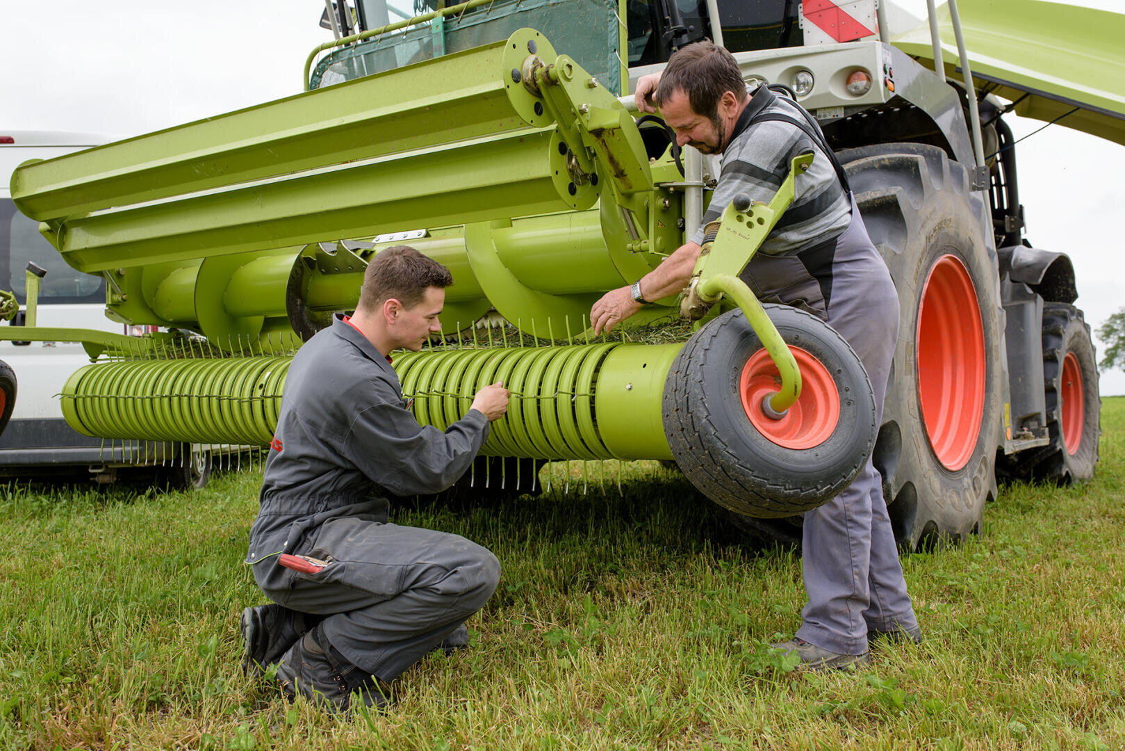 Deux hommes en combinaison de travail réparent ou inspectent la partie avant d'une grande machine agricole verte avec des roues orange, debout dans un champ herbeux.