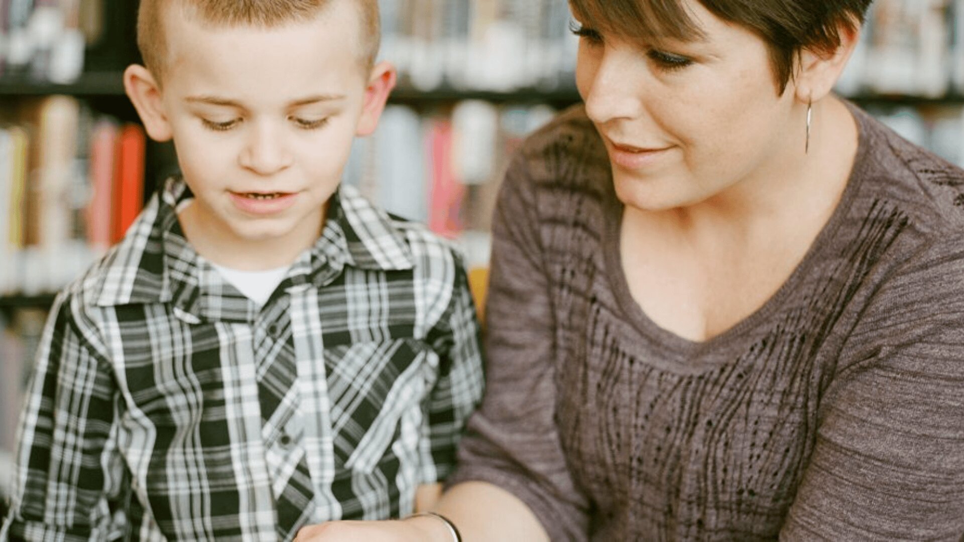 Une femme et un jeune garçon sont assis ensemble devant des étagères et regardent quelque chose devant eux. Le garçon porte une chemise à carreaux et la femme a les cheveux courts et un haut violet.