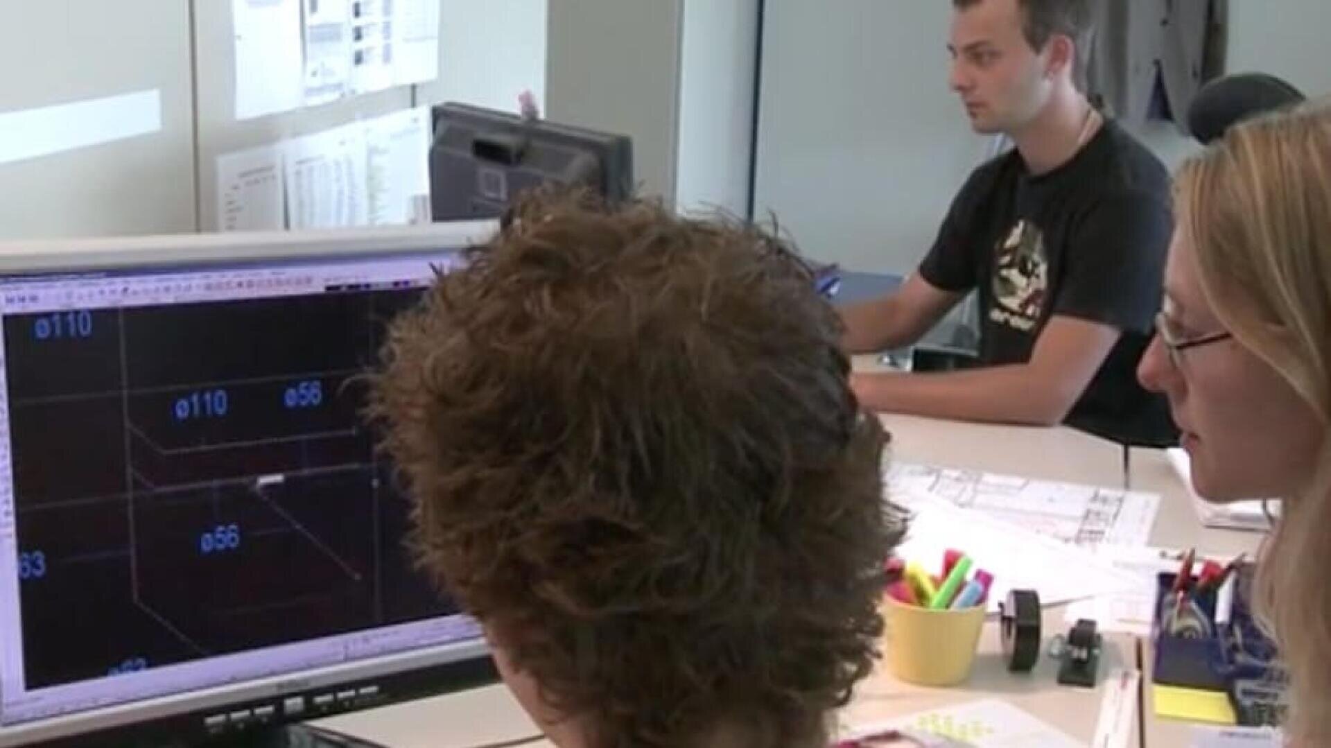 Trois personnes travaillent dans un bureau. Deux d'entre elles regardent un écran sur lequel sont affichés des plans ou des dessins techniques, tandis qu'une troisième personne travaille en arrière-plan. Les bureaux contiennent des papiers, des stylos et des fournitures de bureau.