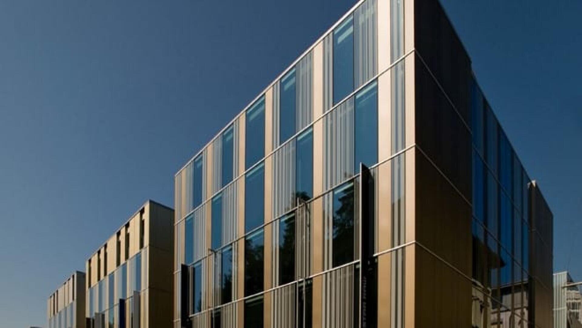Bâtiment moderne doté de grandes baies vitrées et de panneaux verticaux métalliques, reflétant le ciel bleu. La structure présente des lignes épurées et un design minimaliste. La photo est prise en contre-plongée par temps clair.
