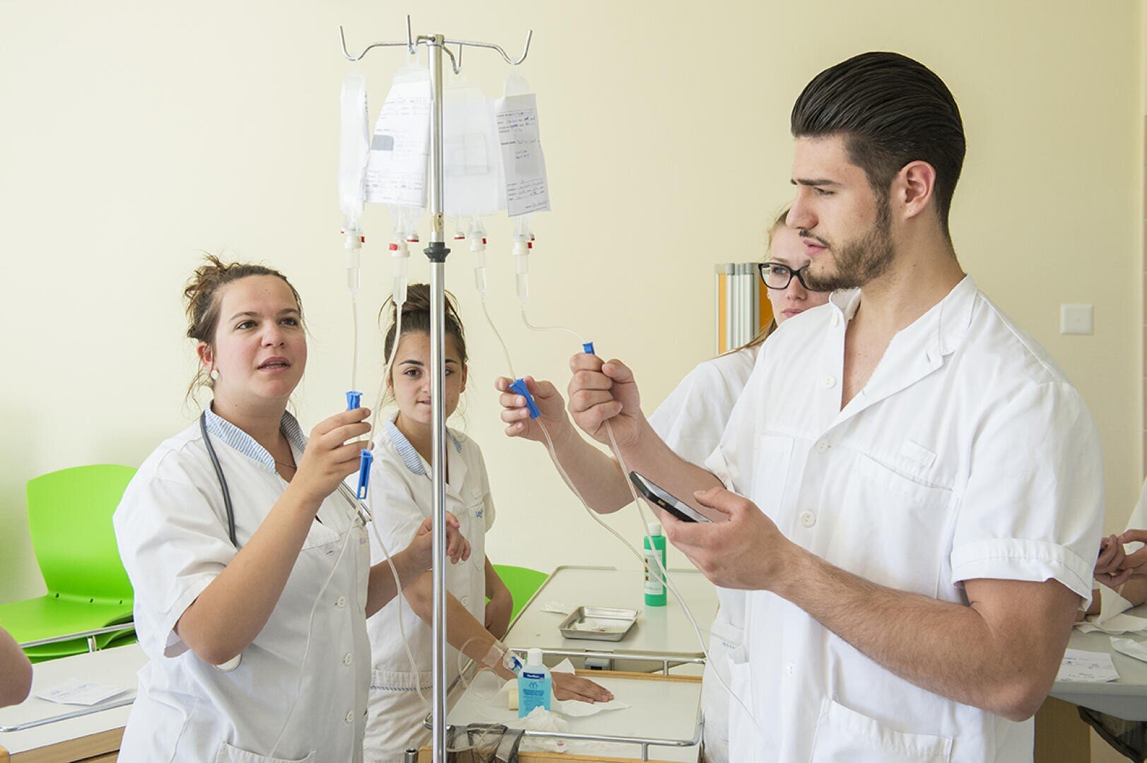 Quatre étudiants en médecine en blouse blanche travaillent ensemble à l'ajustement des poches de liquide intraveineux sur un support dans une salle de classe clinique, semblant concentrés et engagés dans un exercice pratique.