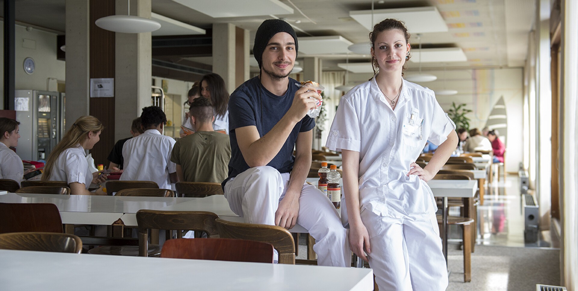 Deux jeunes adultes, l'un en tenue décontractée et l'autre en blouse médicale blanche, posent et sourient dans une cafétéria lumineuse avec d'autres assis et discutant en arrière-plan.