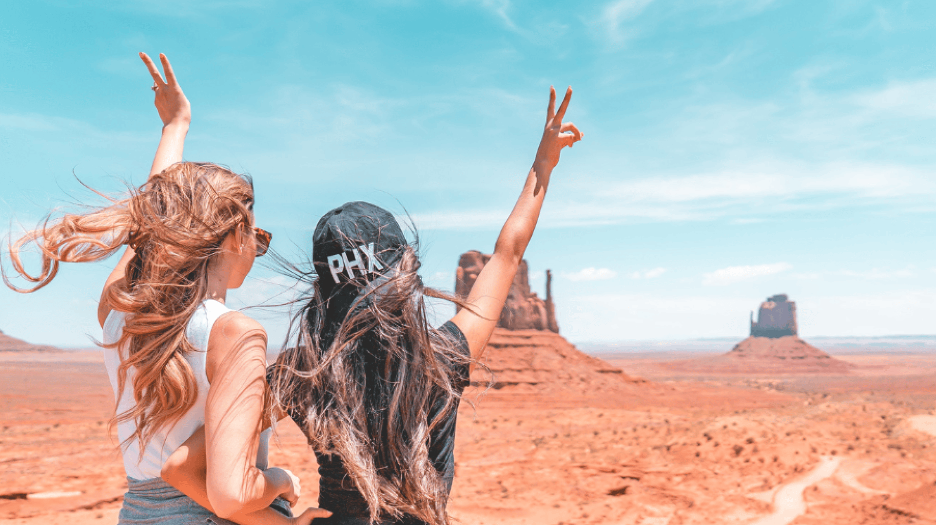 Deux jeunes femmes aux cheveux longs se tiennent dans un paysage désertique, levant les bras et faisant des signes de paix. De grandes formations rocheuses rouges s'élèvent à l'arrière-plan sous un ciel bleu vif. L'une des femmes porte un chapeau PHX noir.
