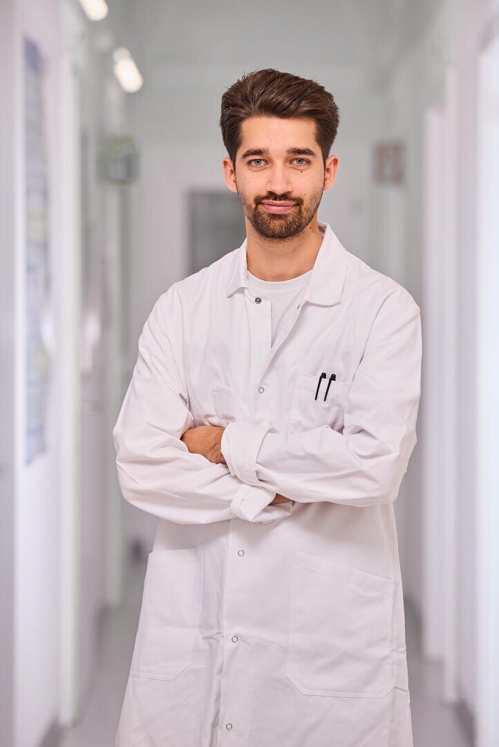 Un jeune homme aux cheveux noirs et à la barbe se tient dans un couloir, vêtu d'une blouse blanche, les bras croisés et souriant à l'appareil photo. L'arrière-plan est légèrement flou, suggérant un environnement clinique ou de laboratoire.