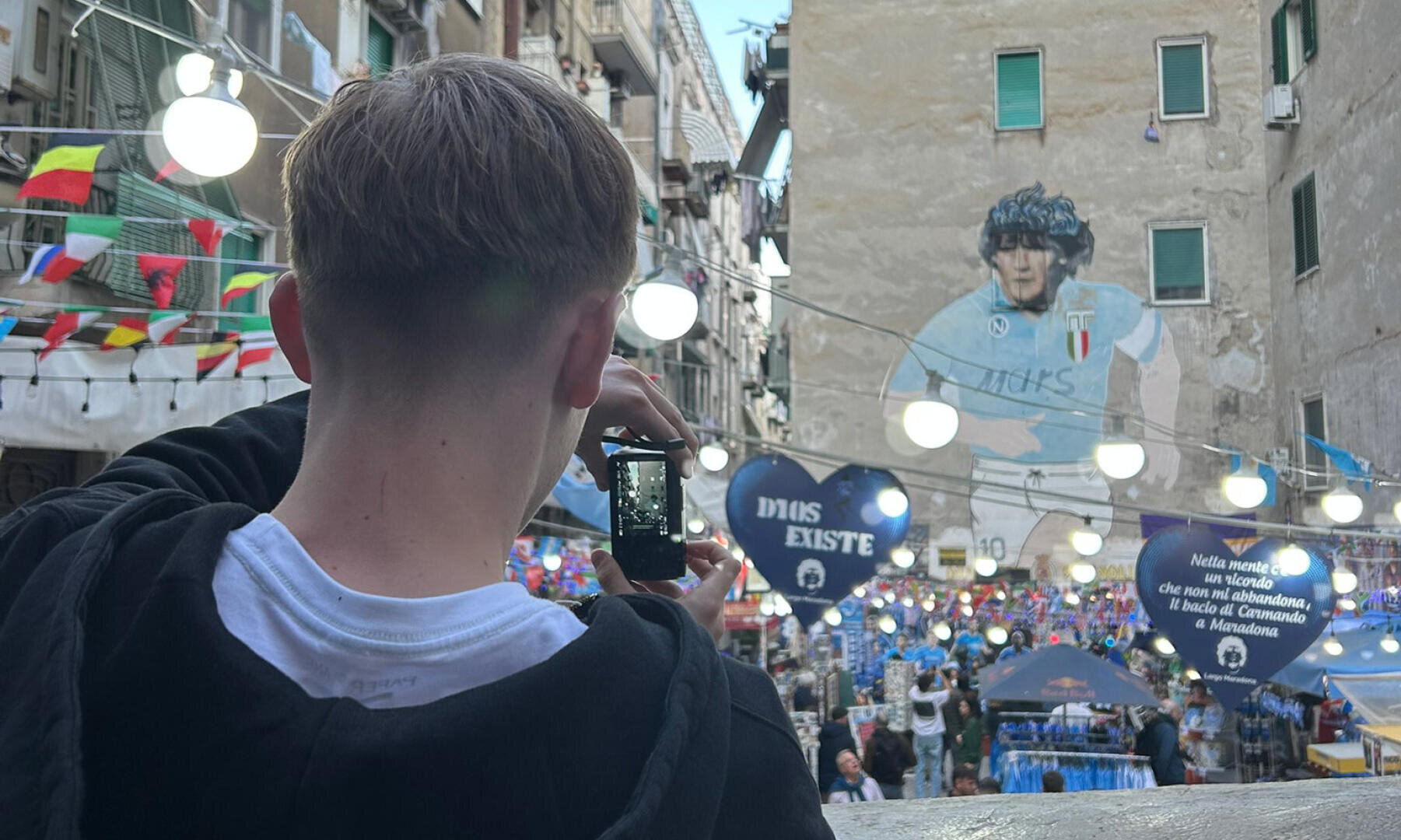 Une personne prend une photo d'une scène de rue animée, avec des décorations et une grande fresque représentant un footballeur sur le mur d'un immeuble. Des drapeaux colorés et des lumières sont suspendus au-dessus de la foule animée en contrebas.