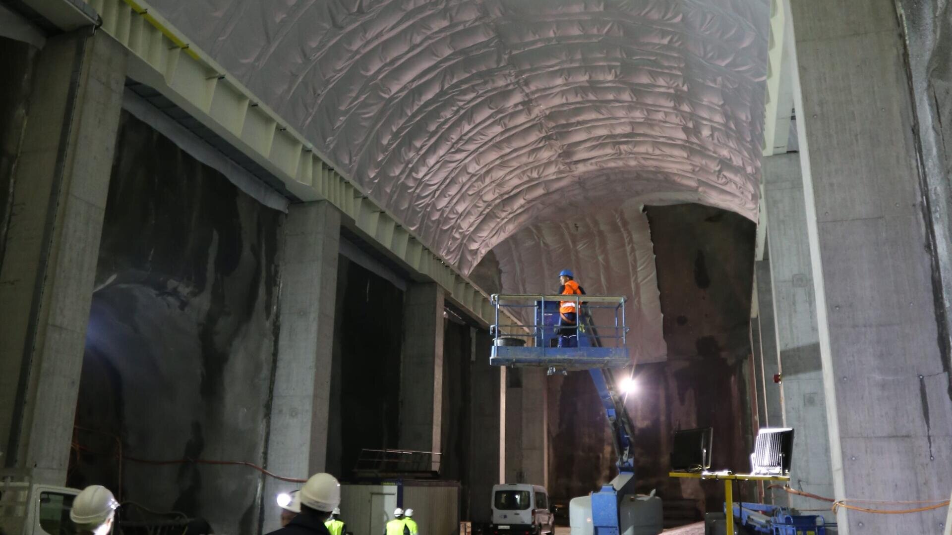 Des ouvriers en tenue de sécurité manœuvrent un élévateur sous un plafond haut, arqué et isolé, à l'intérieur d'un grand tunnel en béton inachevé. Des équipements de construction et des éclairages sont visibles dans tout l'espace.