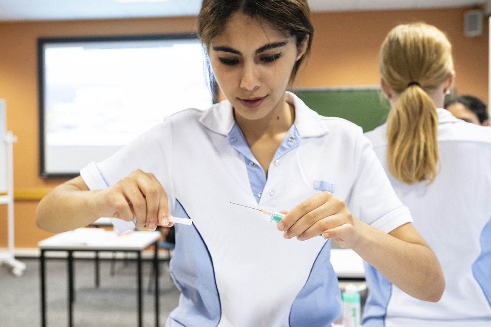 Une jeune femme en uniforme d'infirmière prépare une seringue dans une salle de classe, concentrée sur la manipulation de l'aiguille. D'autres personnes portant des uniformes similaires sont visibles à l'arrière-plan.