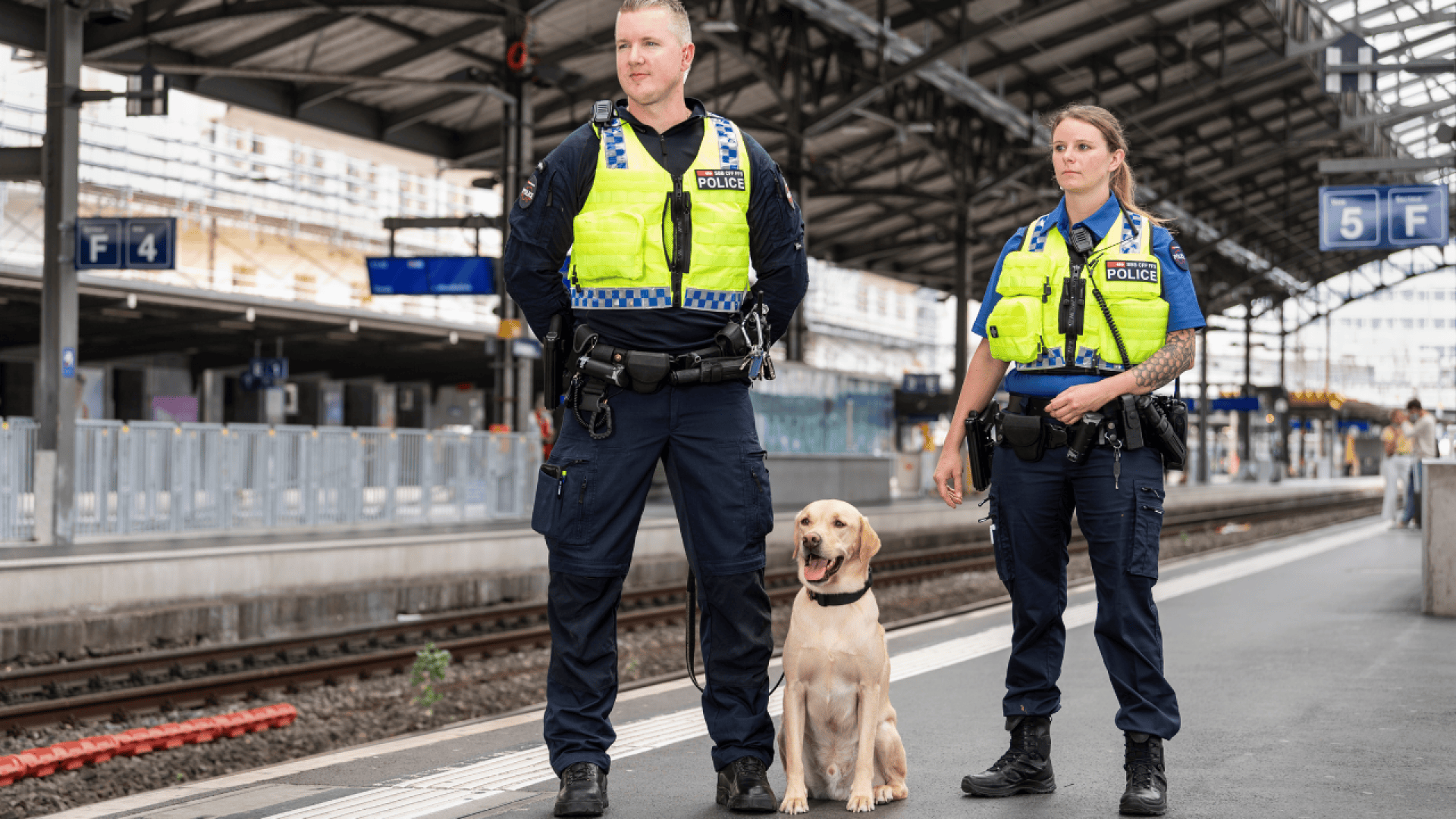 Deux policiers en gilet jaune se tiennent sur un quai de gare avec un labrador jaune. Le quai est vide, les voies et les panneaux de la gare sont visibles à l'arrière-plan.