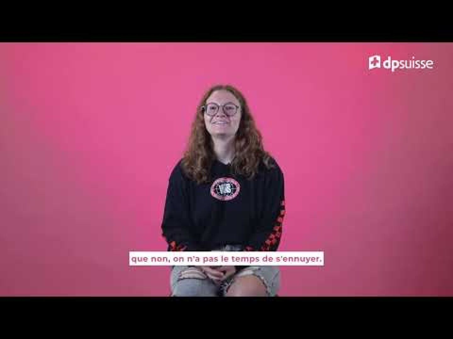 Une jeune femme aux cheveux bouclés et portant des lunettes est assise et sourit devant un fond rose. Elle porte un pull noir orné d'un logo circulaire. Des sous-titres français et le logo dpsuisse apparaissent sur l'image.
