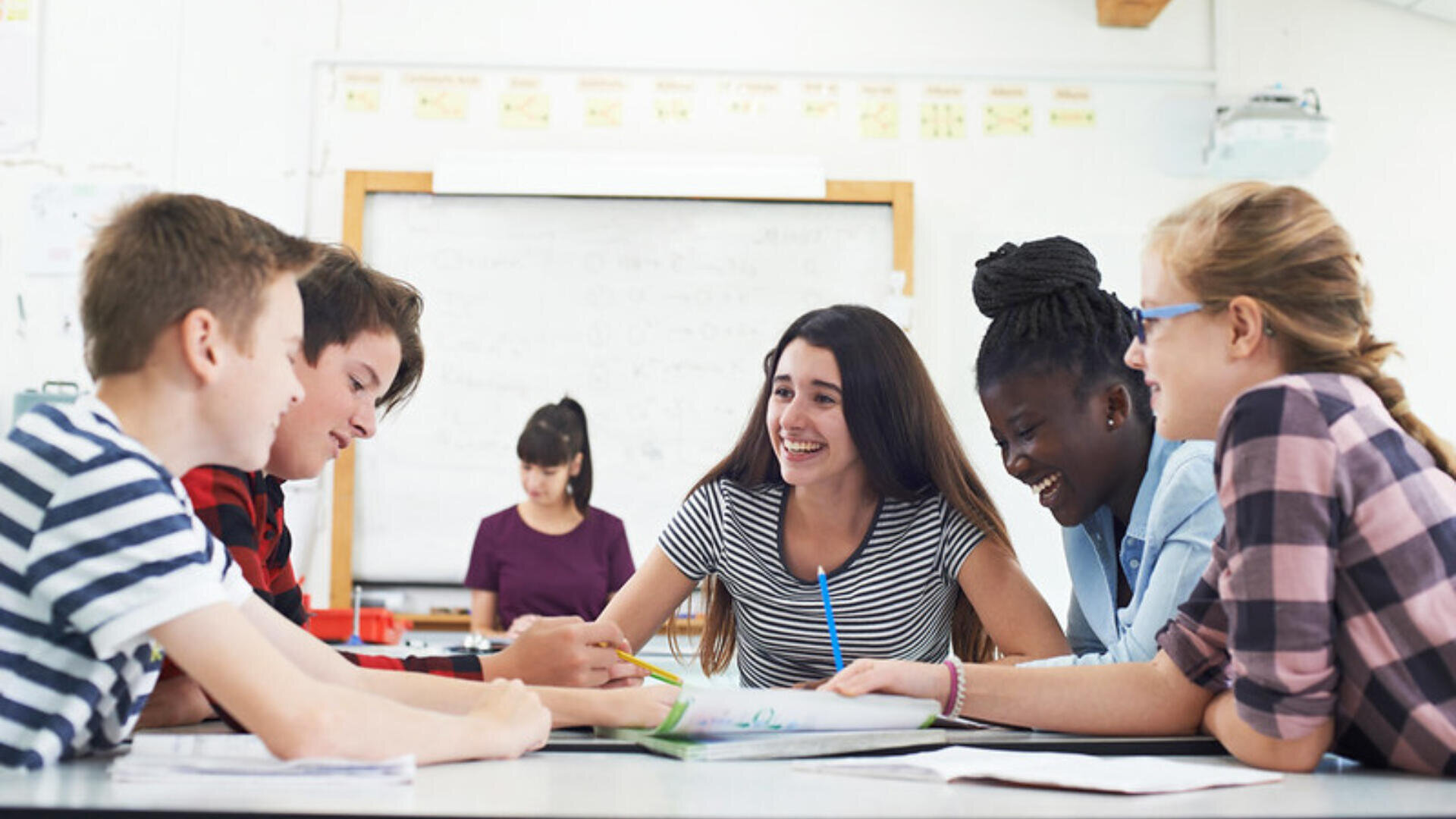 Un groupe de cinq étudiants est assis autour d'une table de classe, souriant et travaillant ensemble sur un projet. Des feuilles et des stylos sont posés sur la table, et un enseignant est visible à l'arrière-plan, près d'un tableau blanc.