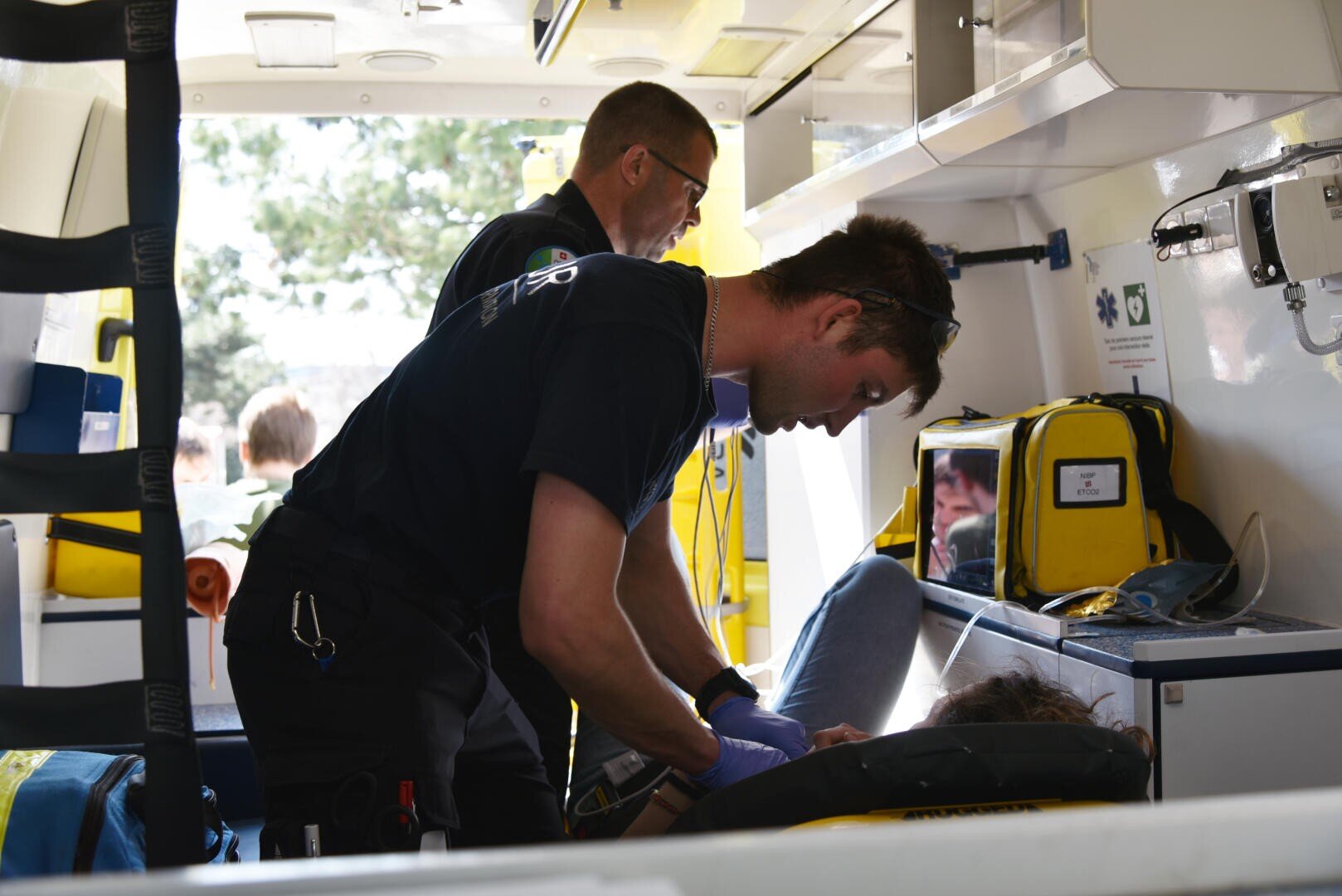 Deux ambulanciers s'occupent d'un patient à l'intérieur d'une ambulance, l'un préparant le matériel médical tandis que l'autre se penche sur le patient sur un brancard. Des sacs et du matériel médical sont visibles autour d'eux.