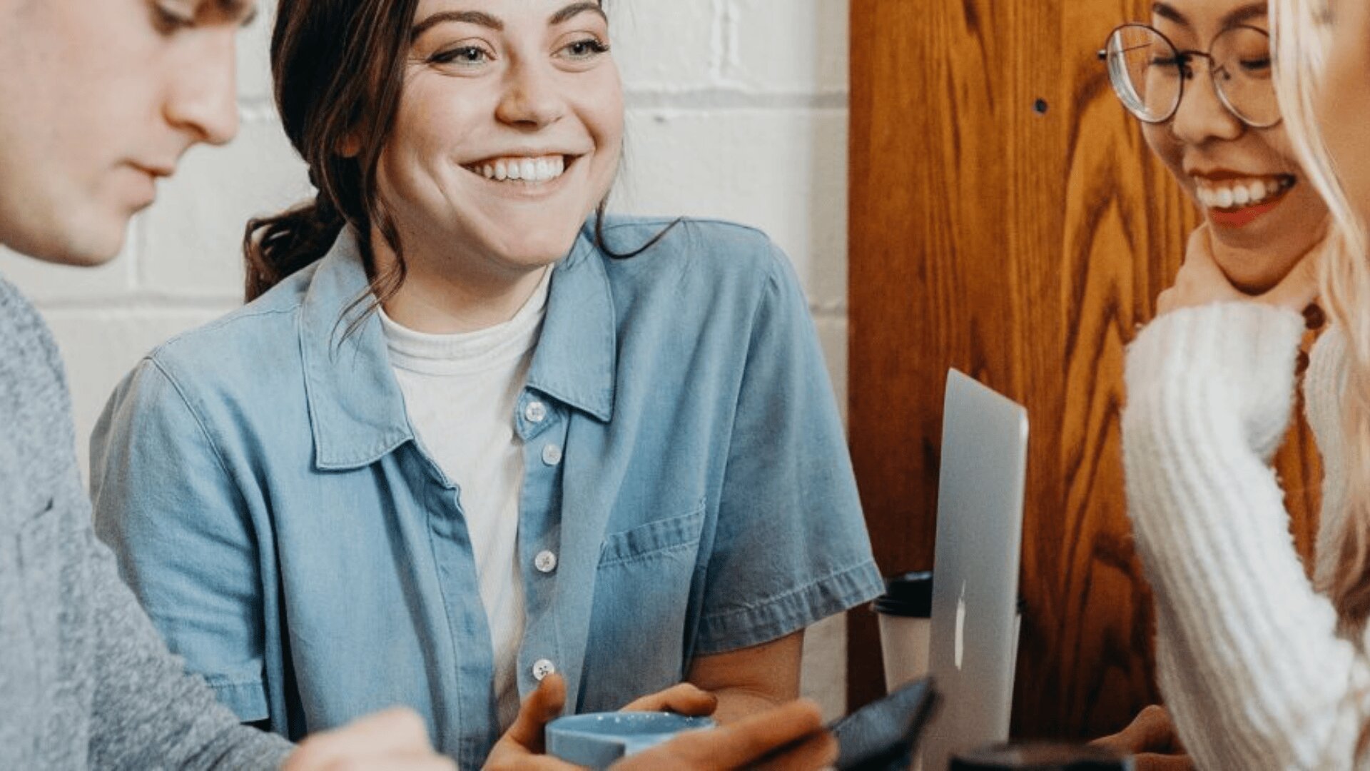 Trois personnes sont assises à une table, souriant et discutant. L'une d'entre elles tient une tasse bleue, un ordinateur portable est ouvert devant elles et elles semblent profiter d'une conversation amicale.