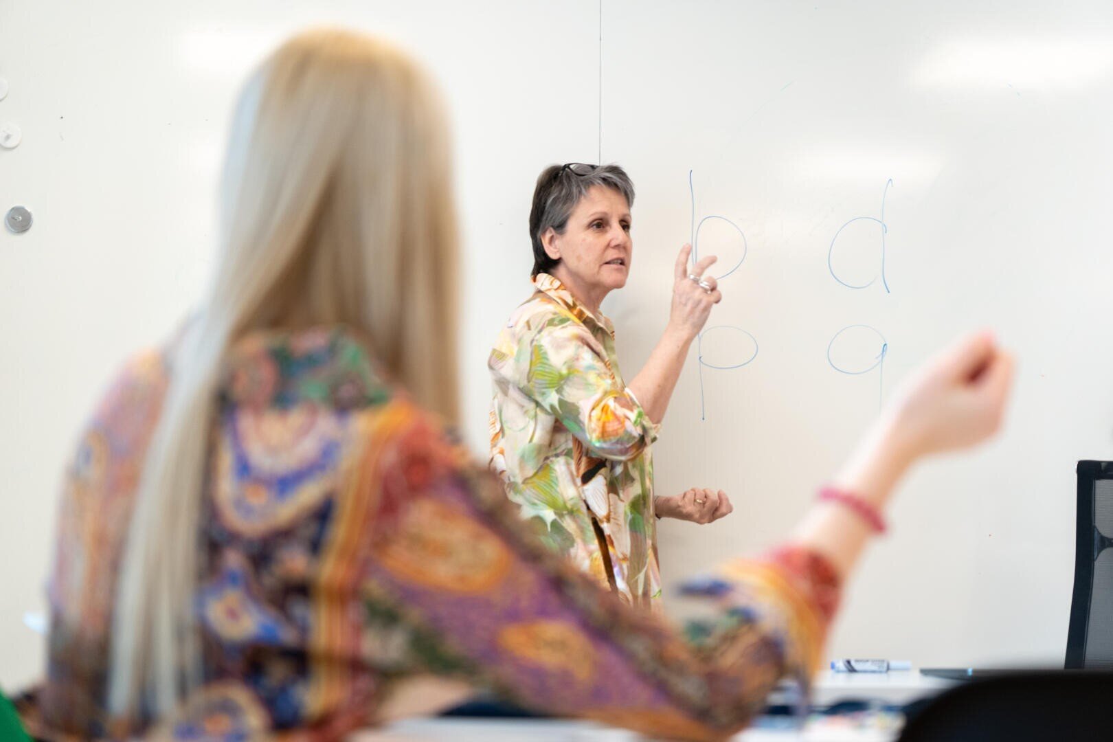 Un enseignant se tient devant un tableau blanc et fait des gestes pour expliquer les lettres qui y sont inscrites (b, d et p). Un élève aux longs cheveux blonds et à la chemise colorée se trouve au premier plan, face à l'enseignant.