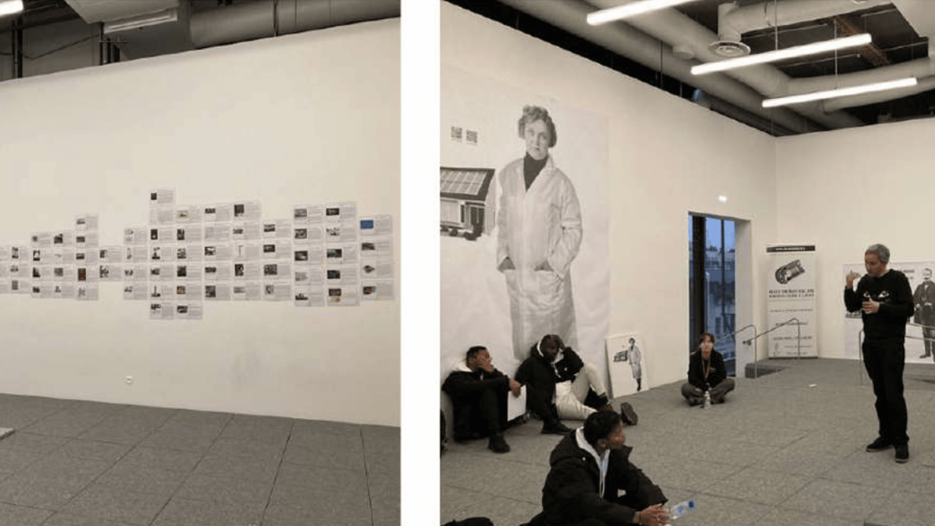 Dans une pièce moderne recouverte d'une moquette grise, des personnes sont assises par terre. Un homme se tient debout et parle. Des documents sont disposés le long d'un mur ; une grande photo en noir et blanc représentant une personne en blouse de laboratoire orne un autre mur.