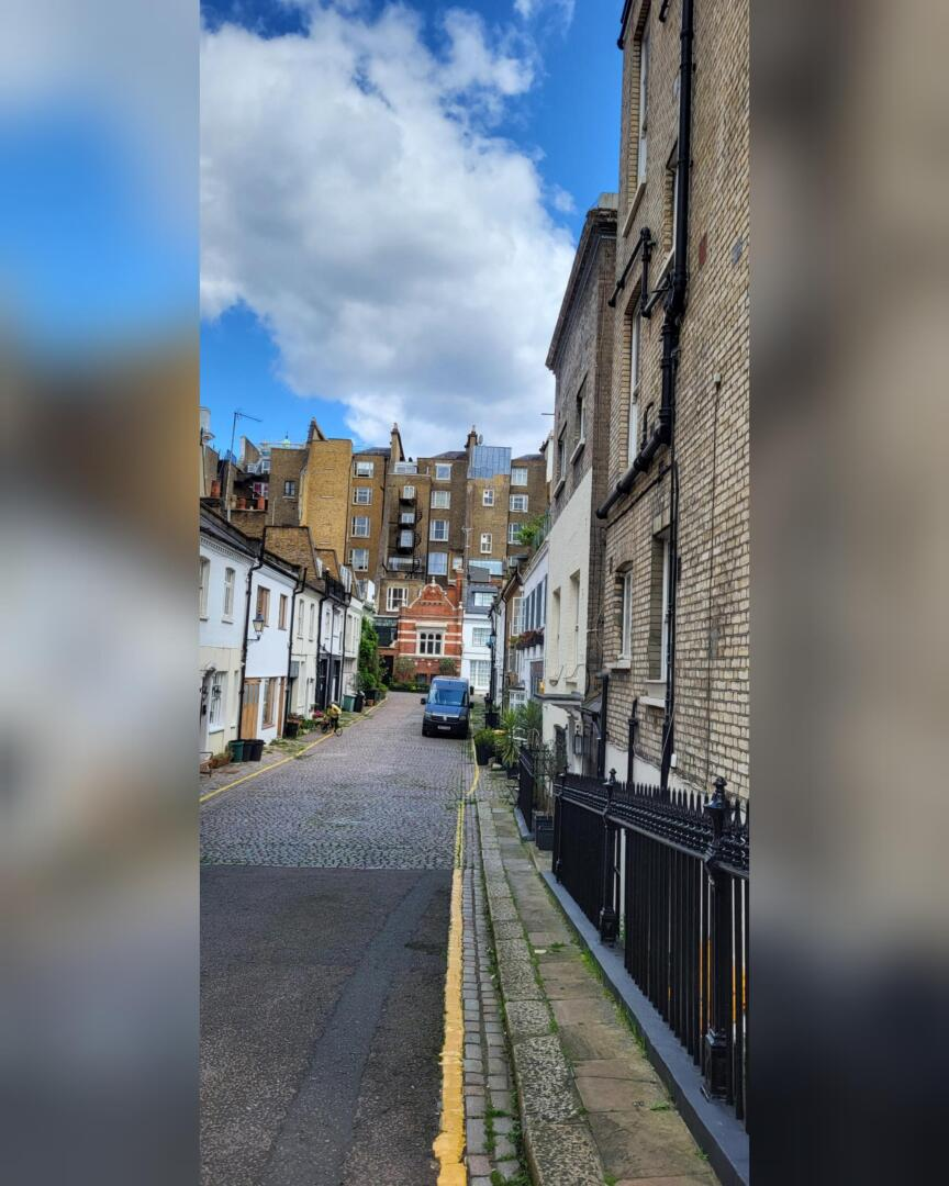 Une rue pavée étroite bordée de part et d'autre de bâtiments blancs et en briques, menant à une camionnette garée au bout ; de grands appartements et un ciel bleu partiellement nuageux sont à l'arrière-plan.