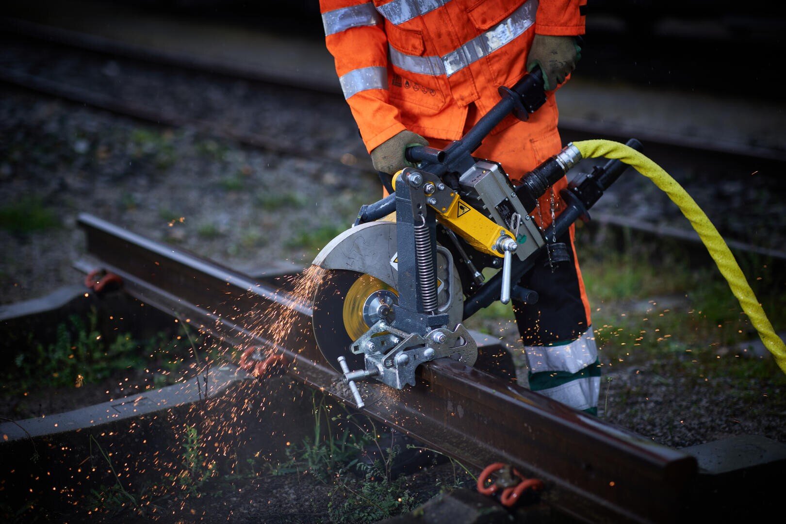Un ouvrier en tenue de signalisation orange coupe une voie métallique avec une tronçonneuse, ce qui provoque des étincelles lumineuses.