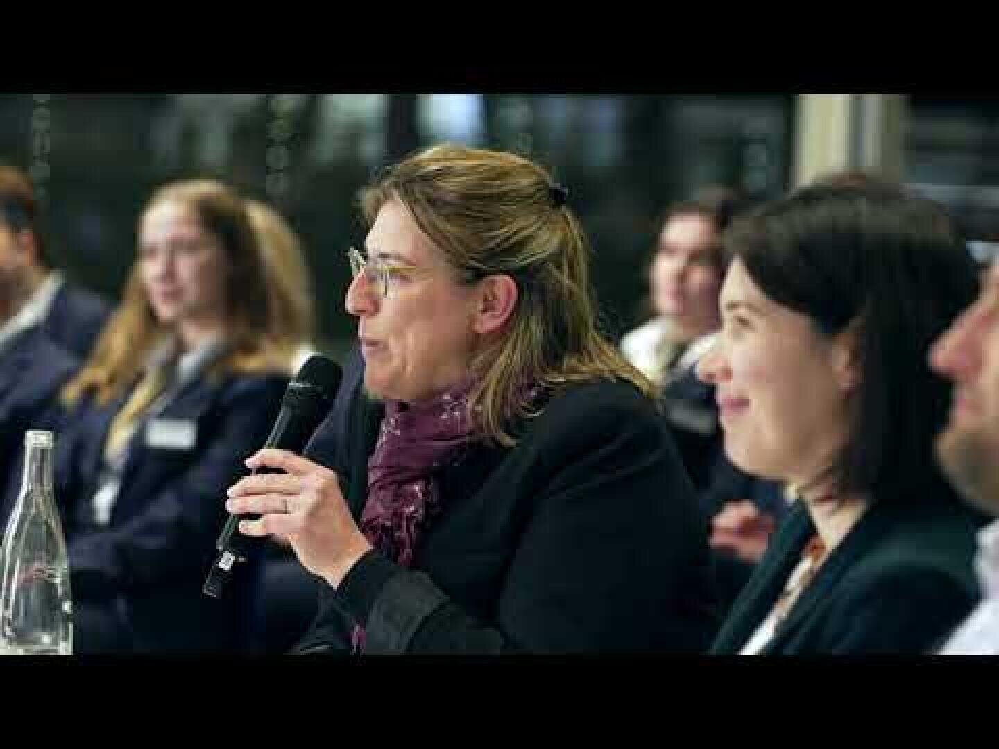 Une femme tenant un micro prend la parole lors d'une table ronde. Plusieurs personnes, hommes et femmes, sont assises à ses côtés et l'écoutent attentivement. Une bouteille d'eau en verre transparent est posée sur la table au premier plan.