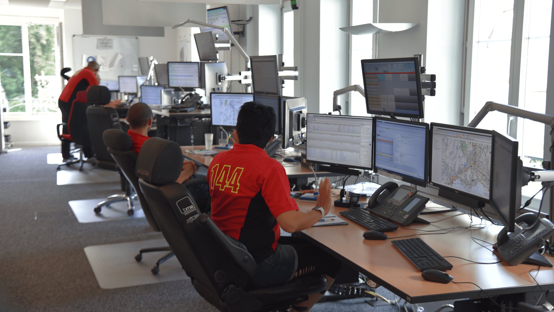 Dans une salle de contrôle, des personnes sont assises à des bureaux équipés de plusieurs écrans d'ordinateur et portent des chemises rouges avec le numéro 144 dans le dos. Ils surveillent les informations et les cartes, et coordonnent probablement les services d'urgence. La lumière du soleil traverse de grandes fenêtres.