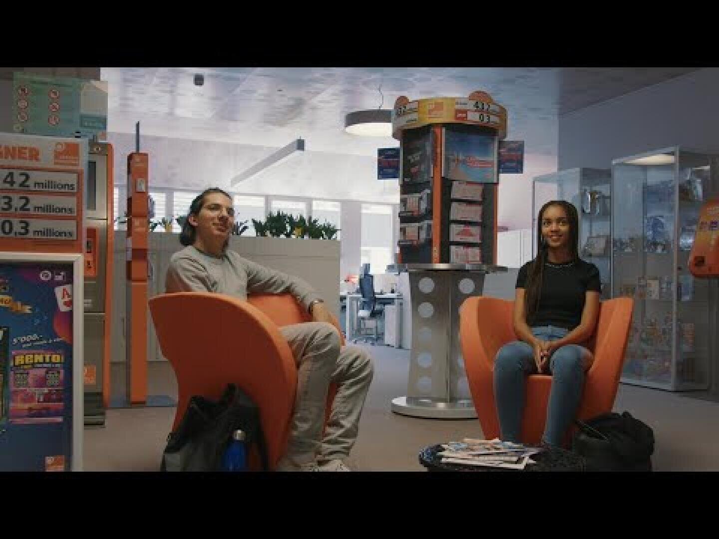 Deux personnes sont assises sur des chaises orange vif dans une salle d'attente moderne et bien éclairée, avec des publicités et des magazines de loterie visibles autour d'elles. Les deux personnes semblent détendues et sont tournées vers l'avant.