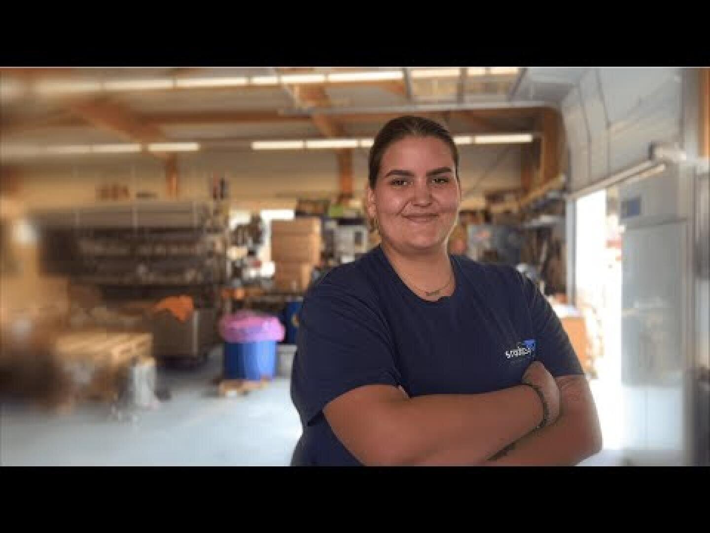 Une personne portant une chemise bleu marine se tient à l'intérieur d'un entrepôt ou d'un atelier, les bras croisés, en souriant à la caméra. L'arrière-plan montre des étagères, des boîtes et des équipements.