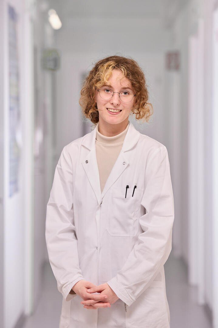 Une jeune femme aux cheveux bouclés et aux lunettes, vêtue d'une blouse blanche et d'un col roulé beige, se tient dans un couloir lumineux en souriant. Deux stylos sont visibles dans la poche de poitrine de sa blouse.