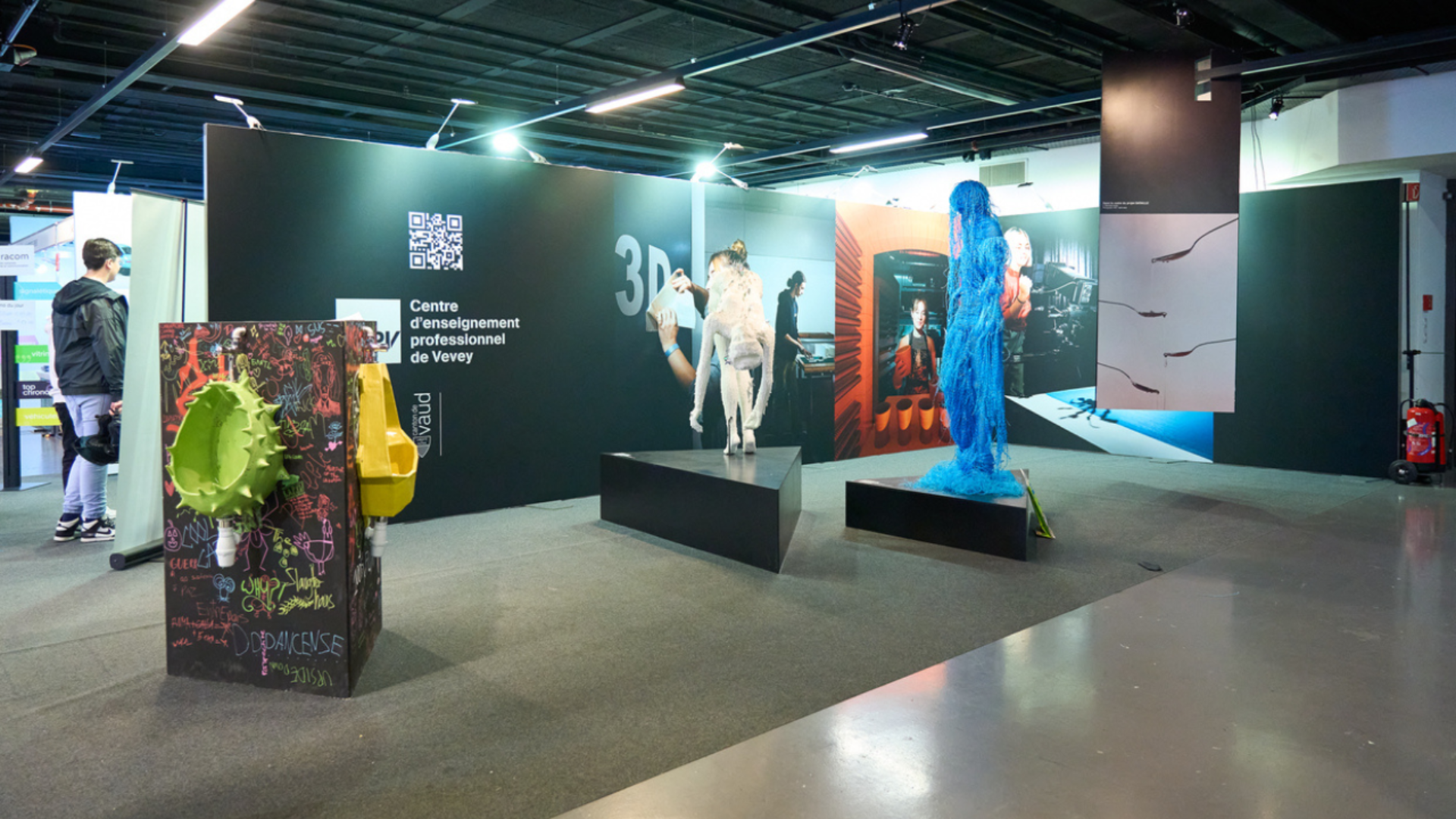 Espace d'exposition d'art avec des mannequins vêtus de costumes créatifs, des installations artistiques colorées et des photos sur des murs d'affichage noirs. Quelques personnes observent les expositions. Le sol et le plafond sont de style industriel.