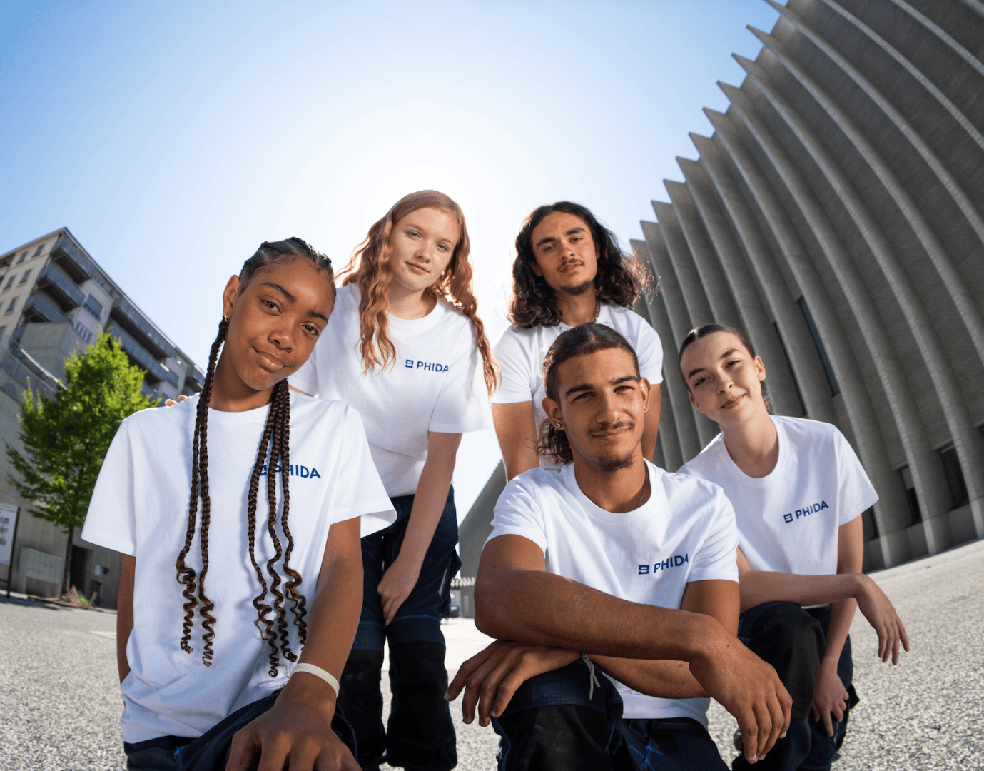 Cinq jeunes vêtus de t-shirts blancs posent ensemble à l'extérieur par une journée ensoleillée, avec des bâtiments modernes en arrière-plan, et regardent l'objectif avec assurance.