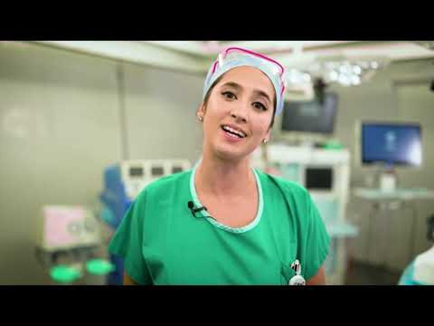 Une femme portant une blouse médicale verte, un bonnet de chirurgien et un micro-casque se tient dans un hôpital ou une salle d'opération très éclairée, regardant la caméra et souriant. Des équipements médicaux sont visibles à l'arrière-plan.