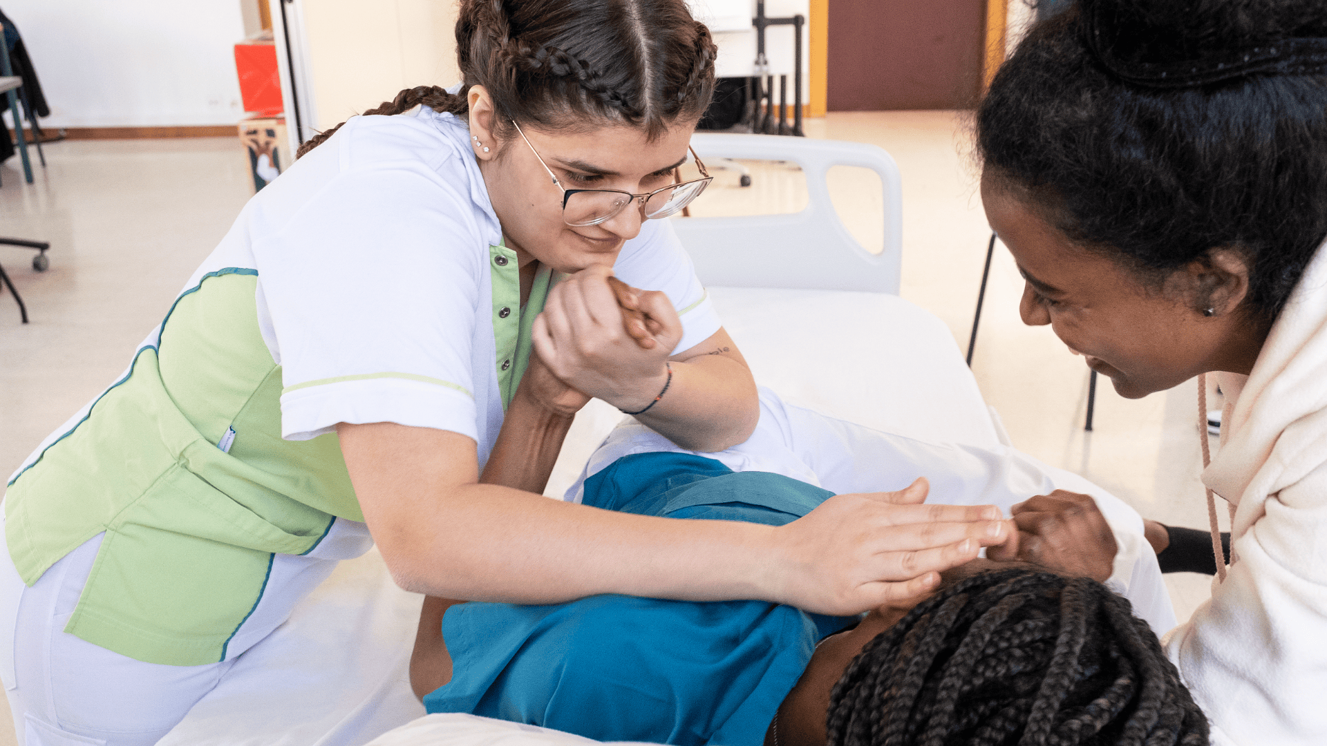 Une infirmière s'occupe d'un patient alité tandis qu'une autre femme se tient à proximité, souriante, lui apportant son soutien. La scène semble se dérouler dans un établissement de santé ou un centre de formation.