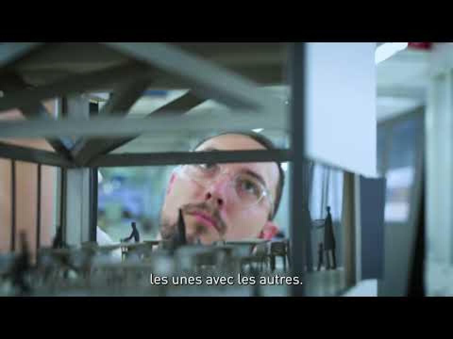 Une personne portant des lunettes examine une maquette architecturale détaillée. En bas de l'écran, on peut lire les sous-titres en français : « les unes avec les autres ».