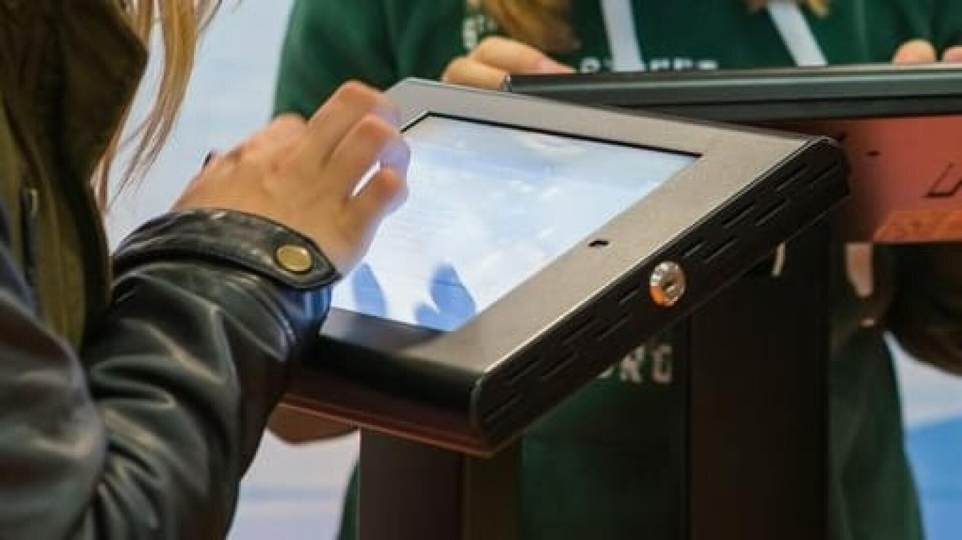 Une personne utilisant un kiosque à écran tactile, vêtue d'une veste sombre. Une autre personne portant un sweat à capuche vert est partiellement visible à l'arrière-plan.