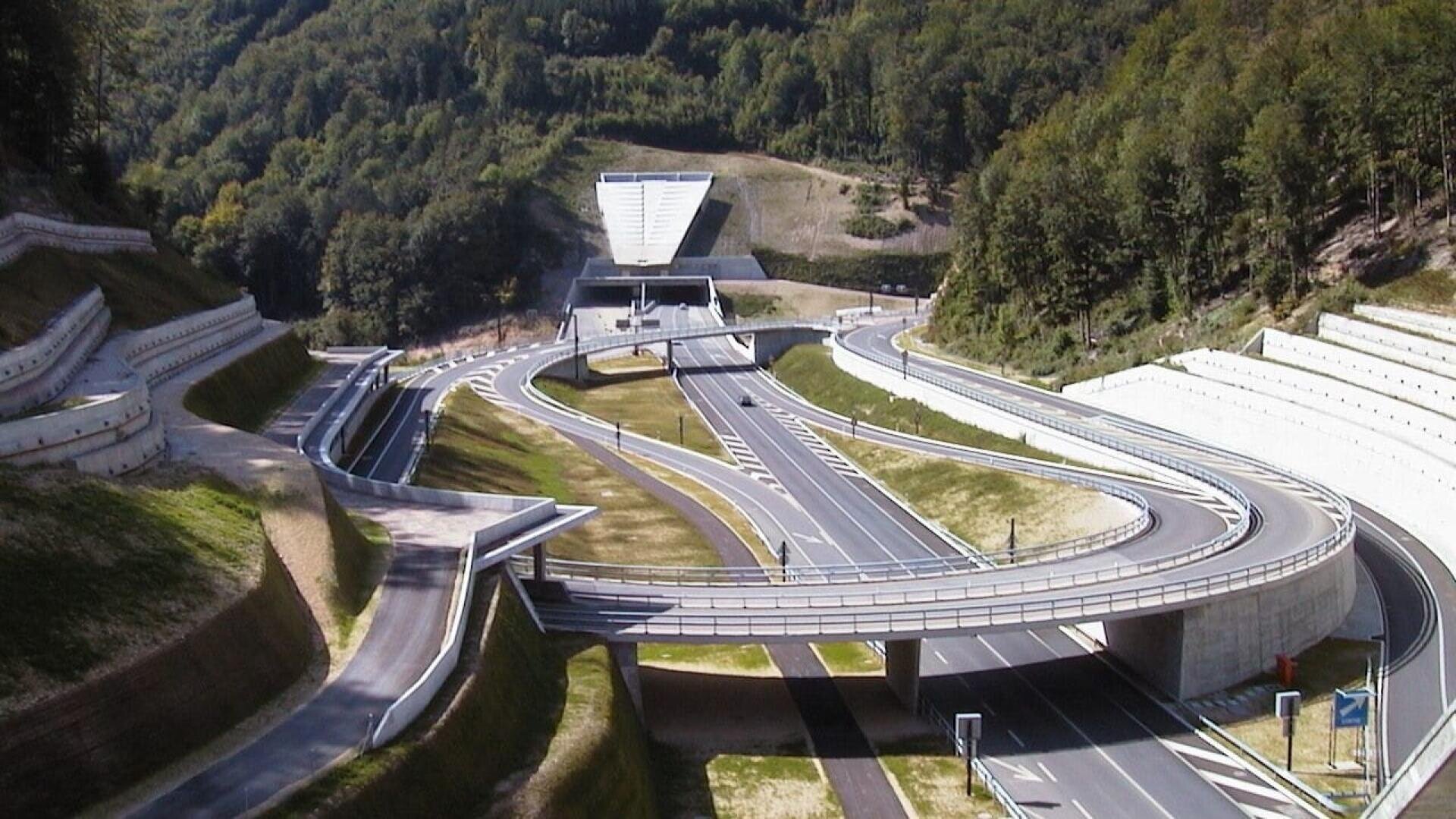 Un échangeur autoroutier moderne avec de multiples rampes surélevées et des routes courbes mène à l'entrée d'un tunnel entouré de collines verdoyantes et boisées sous un ciel dégagé.
