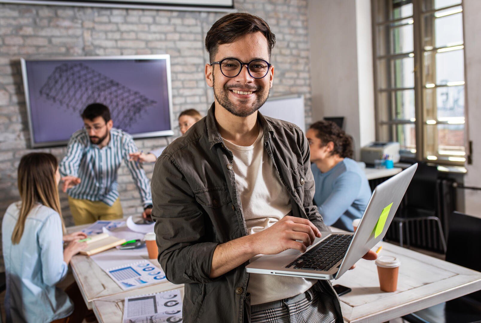 Un homme souriant tient un ordinateur portable dans un bureau moderne tandis que quatre collègues collaborent à une table à l'arrière-plan. Des tableaux et des documents sont étalés sur la table, et une présentation est affichée sur un écran.