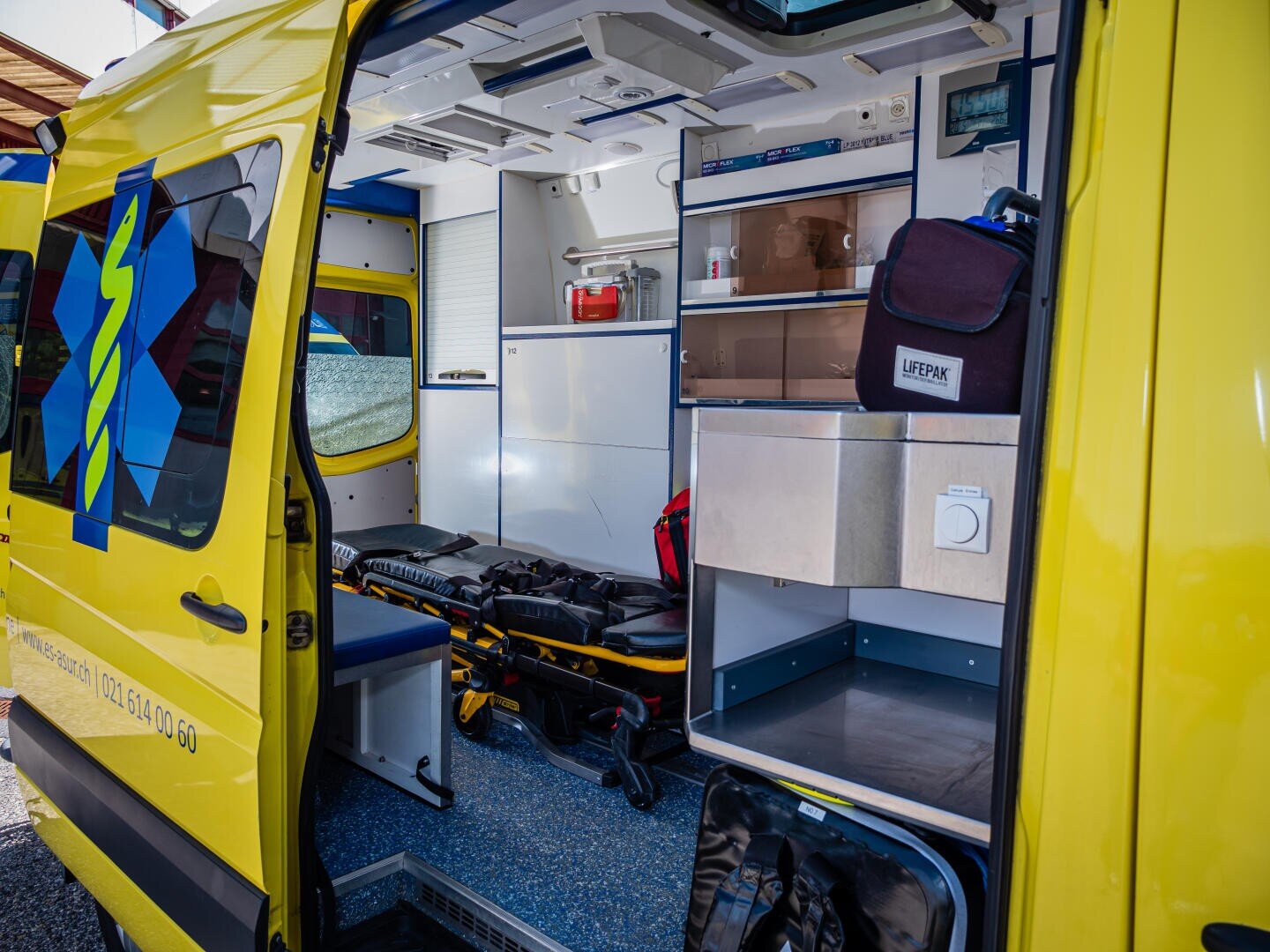 L'image montre l'intérieur d'une ambulance jaune dont la porte latérale est ouverte, révélant du matériel médical, un brancard, des sièges, des compartiments de rangement et un sac LIFEPAK.