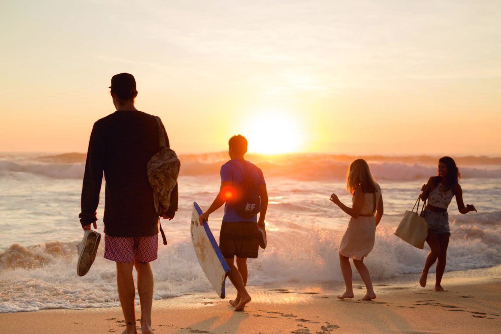 Quatre personnes marchent vers l'océan au coucher du soleil, portant des sacs de plage et du matériel de surf. Les vagues s'écrasent sur le rivage et le soleil est bas sur l'horizon, créant une lueur chaude et vibrante sur la scène.