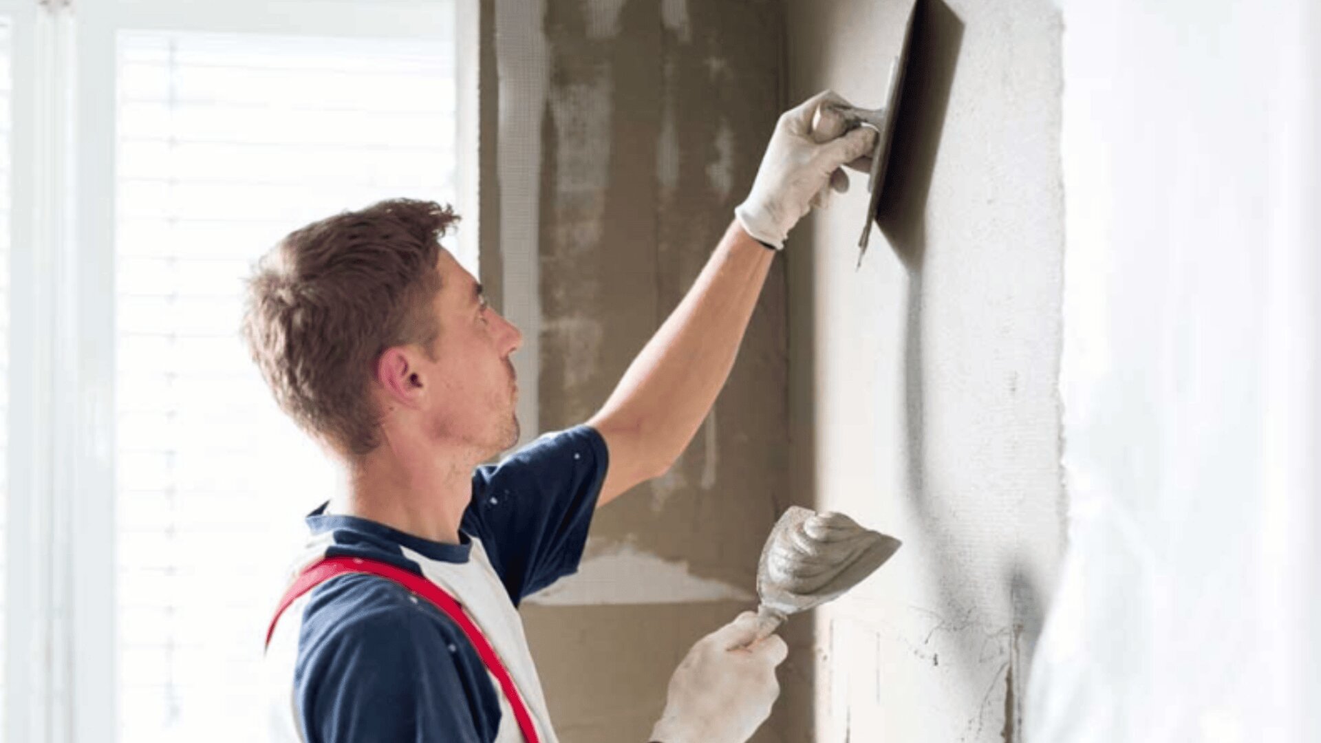 Un homme portant des gants de travail et une chemise blanche avec des bretelles rouges lisse le plâtre sur un mur à l'aide d'une truelle dans une main et tient plus de plâtre sur un faucon dans l'autre main.