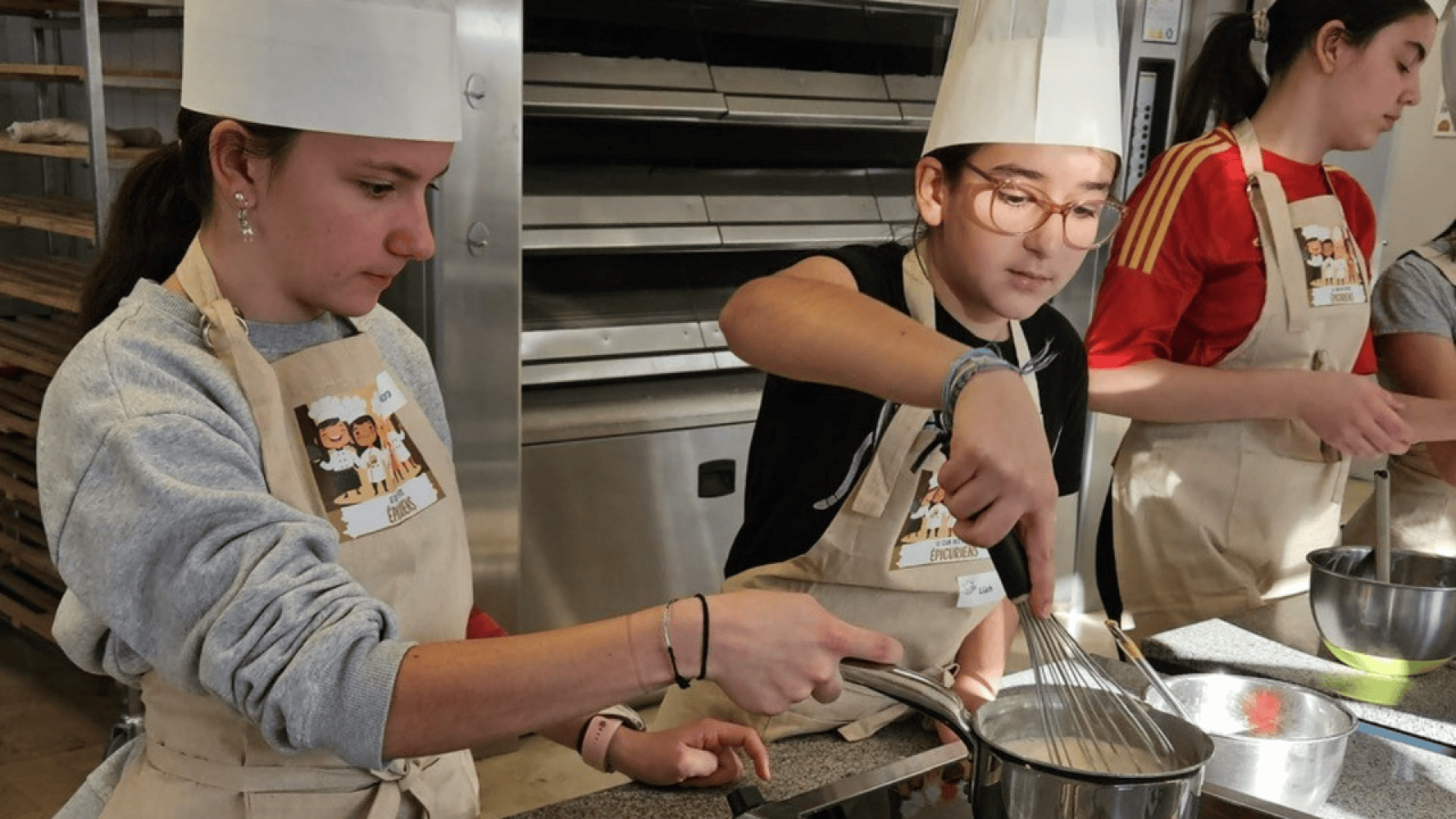 Deux jeunes gens portant des toques et des tabliers cuisinent ensemble, fouettant des ingrédients dans une casserole sur le comptoir d'une cuisine, tandis qu'une autre personne prépare des aliments à l'arrière-plan.