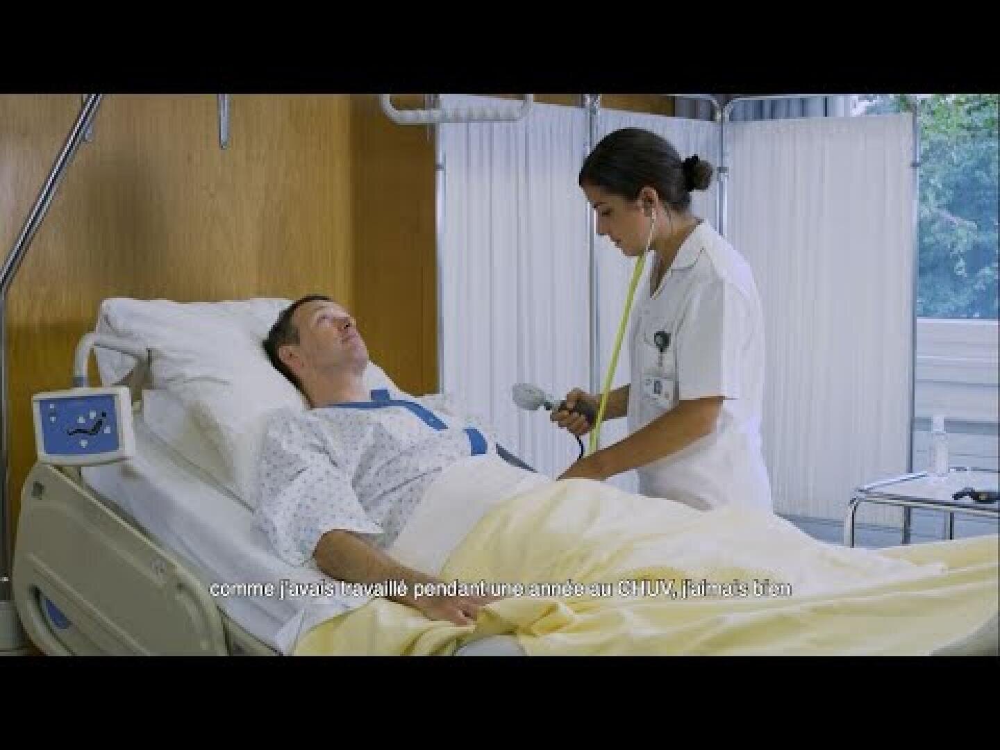 Une infirmière se tient à côté d'un lit d'hôpital et s'occupe d'un patient allongé avec une couverture jaune. Du matériel médical est visible et un texte en français apparaît au bas de l'image.