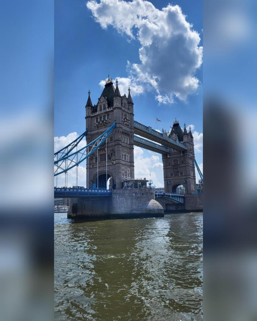 L'image montre le Tower Bridge de Londres par une journée ensoleillée, avec un ciel bleu et des nuages au-dessus. Ce pont emblématique enjambe la Tamise et ses tours jumelles s'élèvent au-dessus de l'eau, se reflétant doucement à la surface.